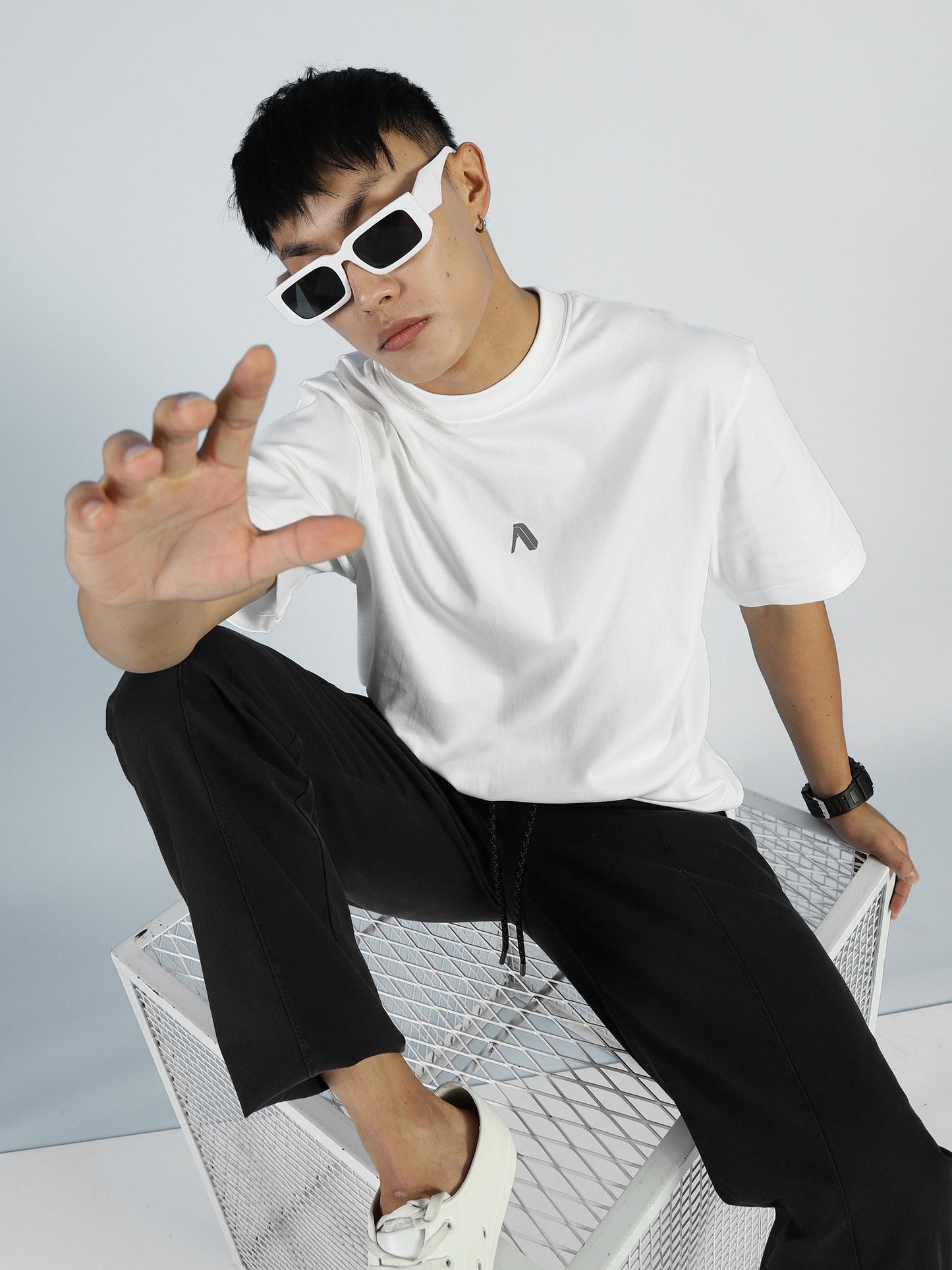 A modern trailblazer sports My Store’s Vibe T in Bright White, white sunglasses, black pants, and white sneakers while sitting on a metal mesh cube, reaching towards the camera against a light background.